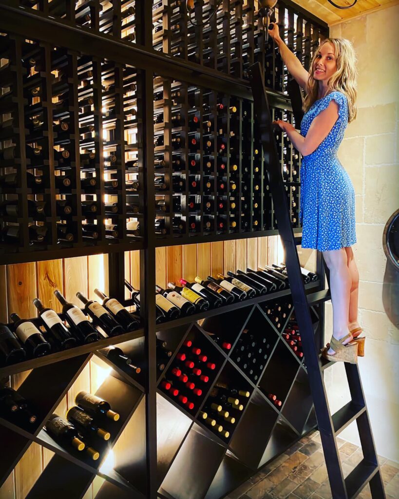 Woman in wine cellar reaching for wine bottle.